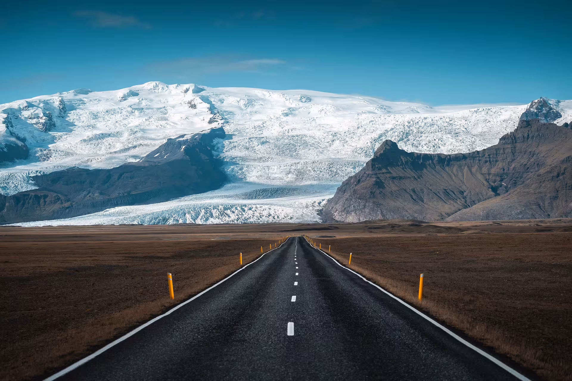 Straight road leading toward Vatnajökull glacier and snow-capped mountains in Iceland under blue sky
