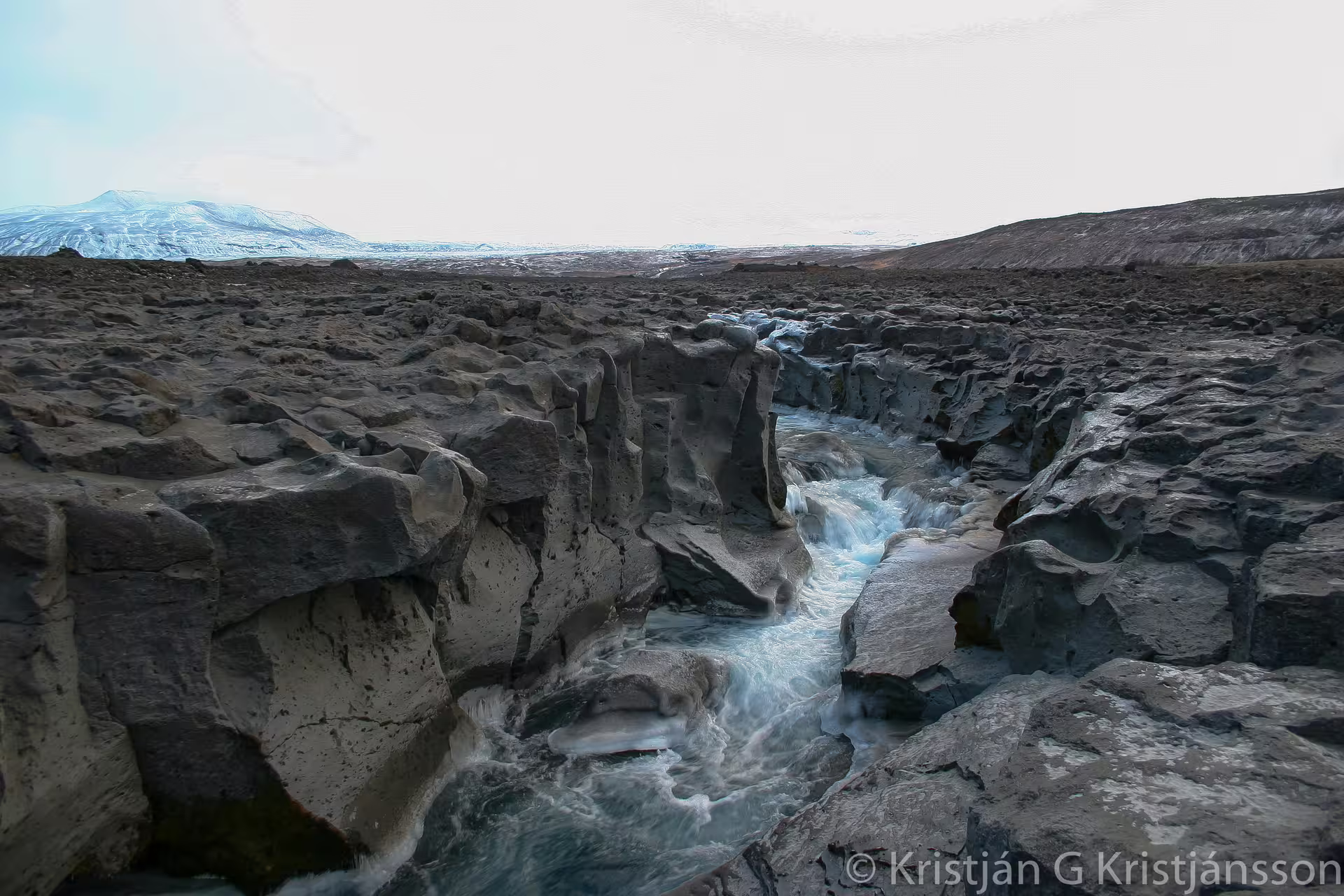 Glacial meltwater rushing through a rocky lava canyon with snow-capped mountains in Iceland