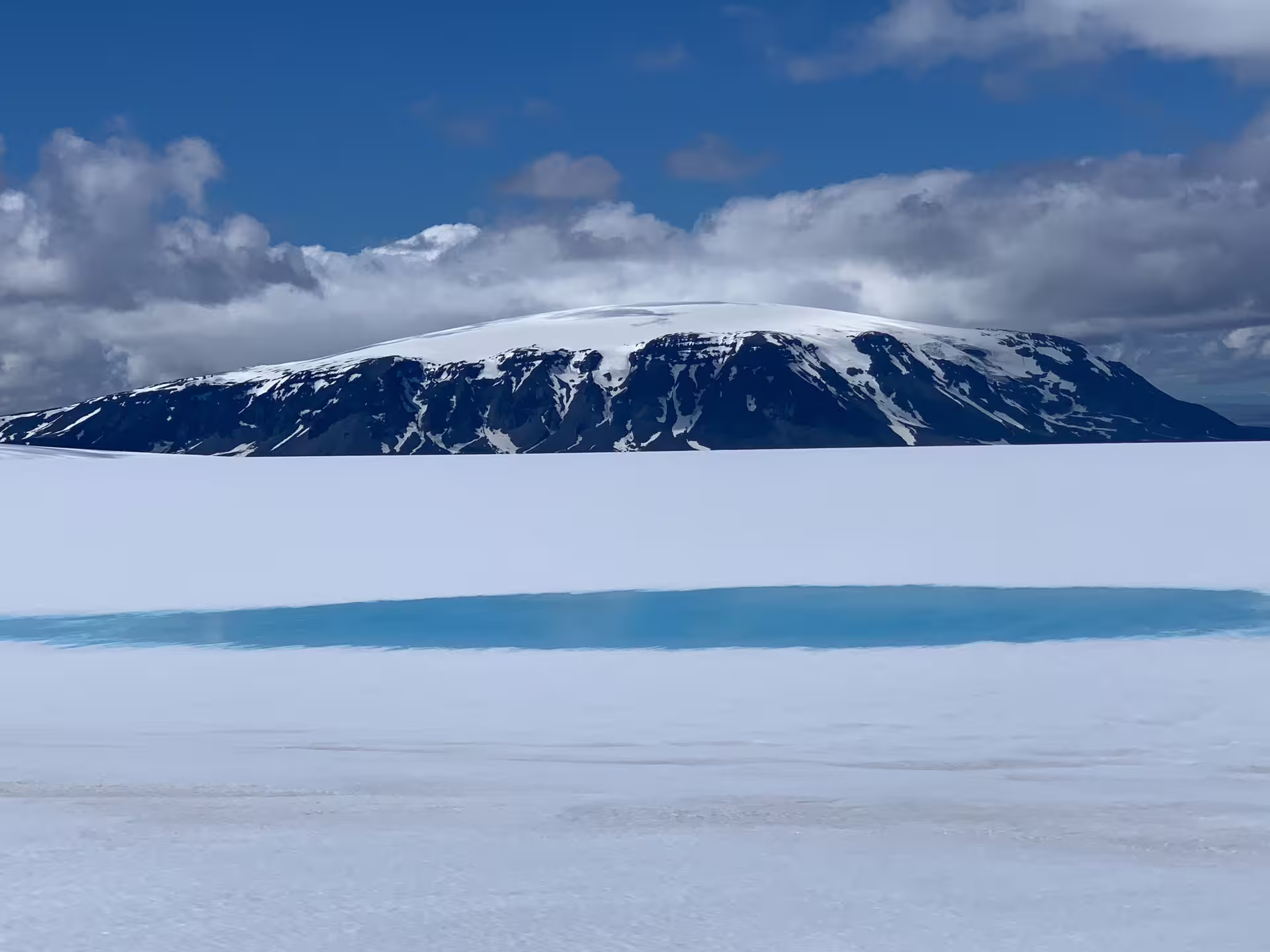 Snow-covered glacier with volcanic mountain and turquoise meltwater pool under blue sky