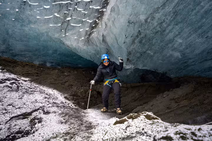 Guide posing inside Solheimajokull glacier cave wearing blue helmet and crampons, holding ice axe