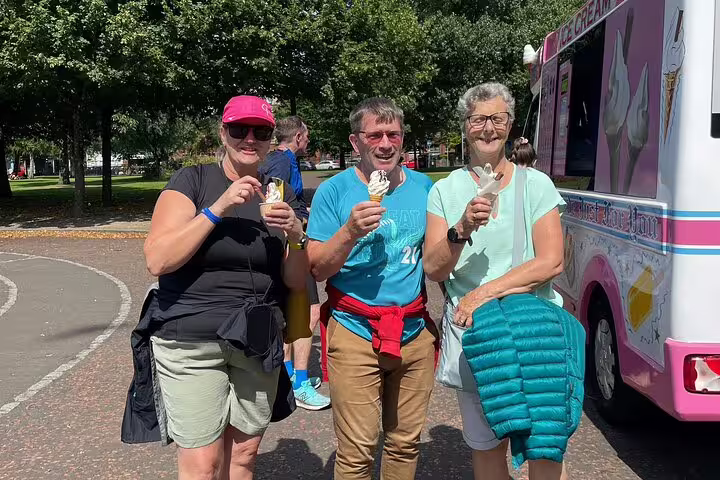 Three smiling people enjoying ice cream cones near a pink ice cream truck in a sunny Glasgow park