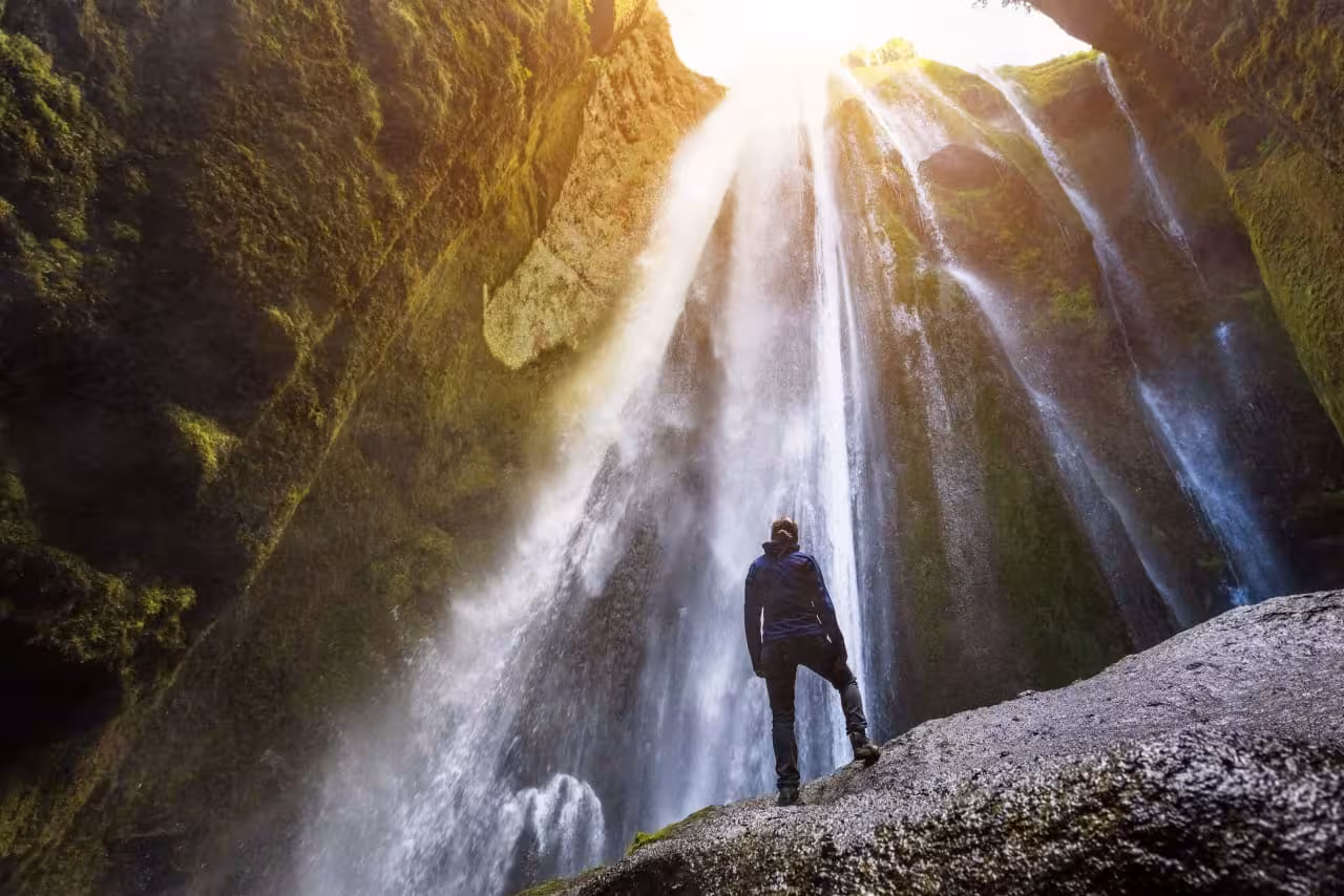 Hiker stands in awe beneath Gljufrabui hidden waterfall with sunrays on Iceland South Coast full-day tour