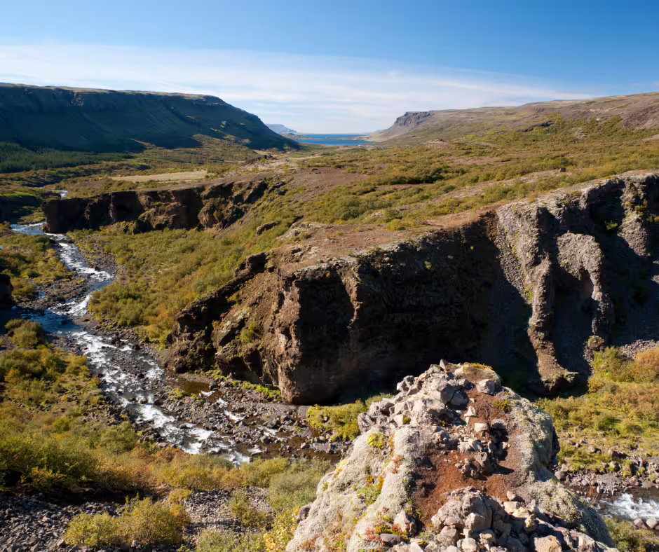 Rocky canyon with a rushing river winding through green valley toward a fjord under blue skies near Glymur Iceland