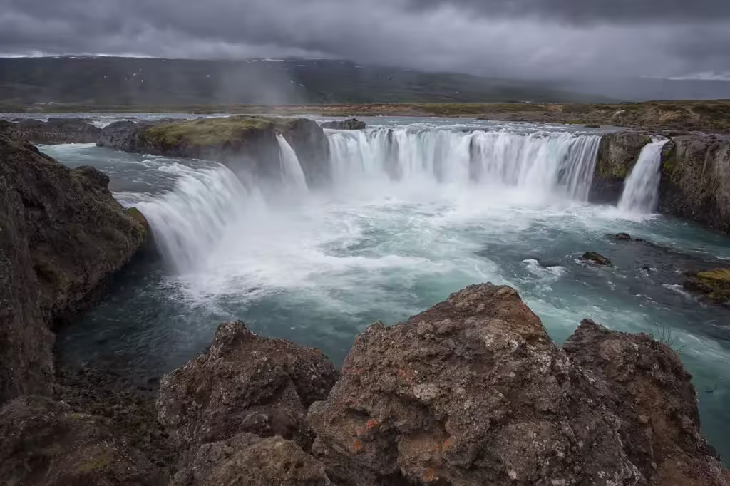 Godafoss waterfall cascading over wide basalt cliffs into turquoise waters under dramatic cloudy skies, Iceland