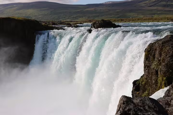 Godafoss waterfall cascading over wide rocky ledge with misty spray and green hills in background, Iceland