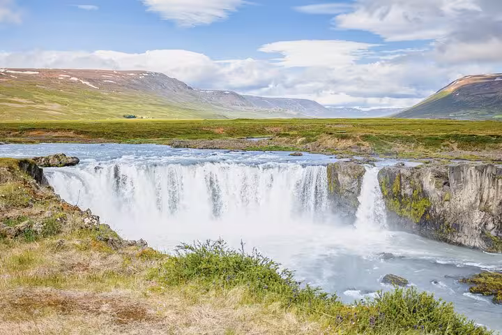 Godafoss waterfall cascading over rocky cliffs with green meadows and snow-capped mountains near Akureyri, Iceland