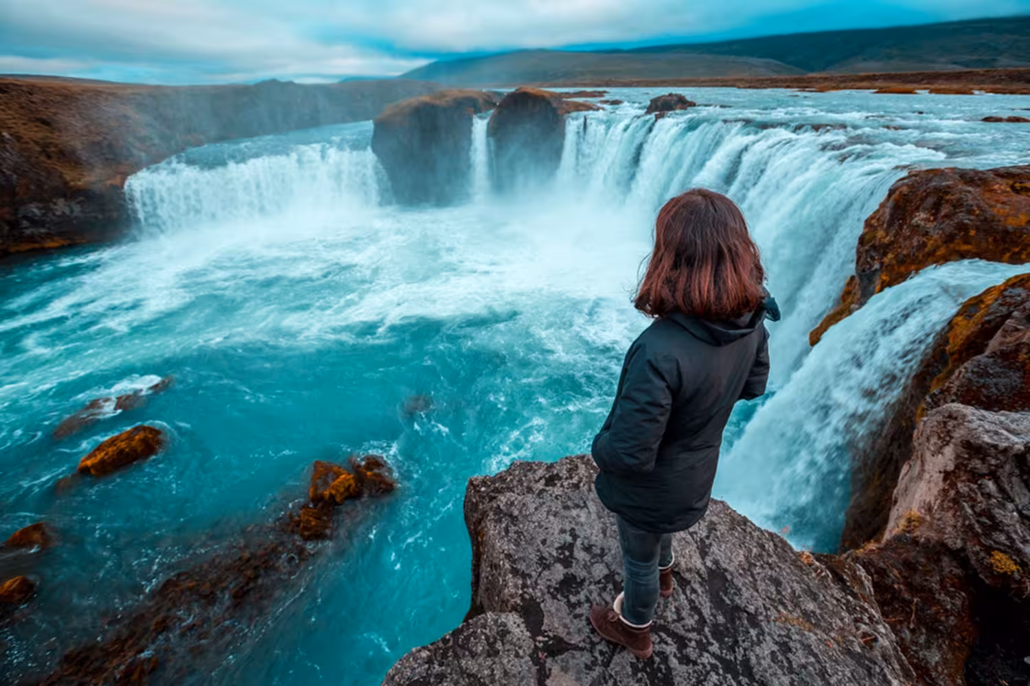 Woman standing on rocky cliff overlooking Godafoss waterfall with turquoise waters in Iceland