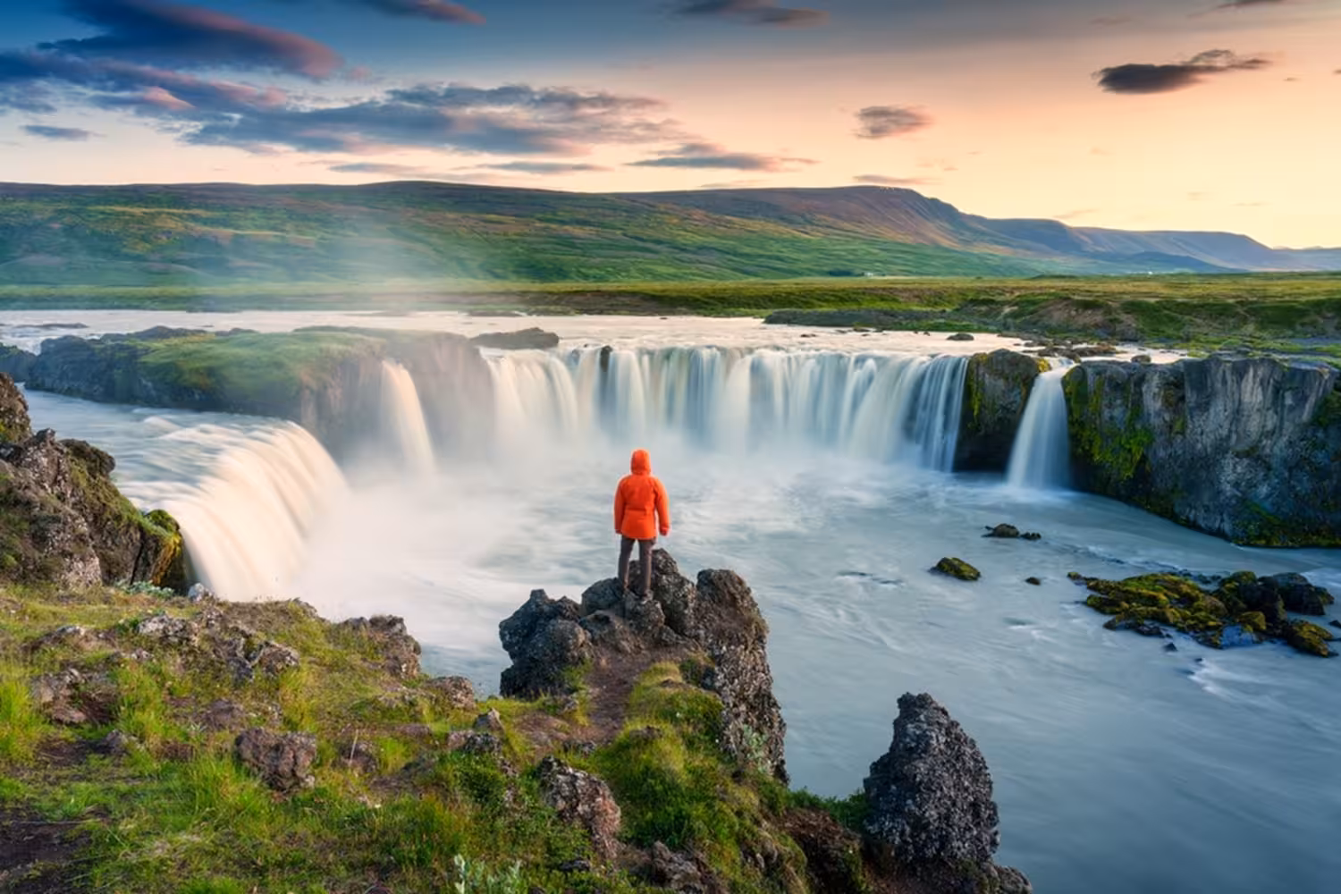 Person in red jacket standing on rocks overlooking Godafoss waterfall in Iceland