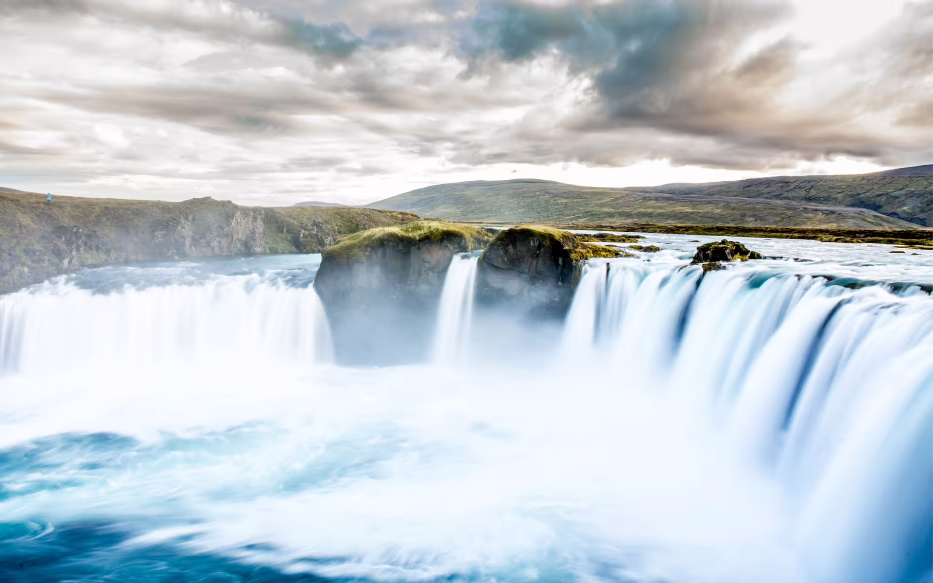 Godafoss waterfall cascading over rocky ledges with misty blue water and rolling hills in Iceland