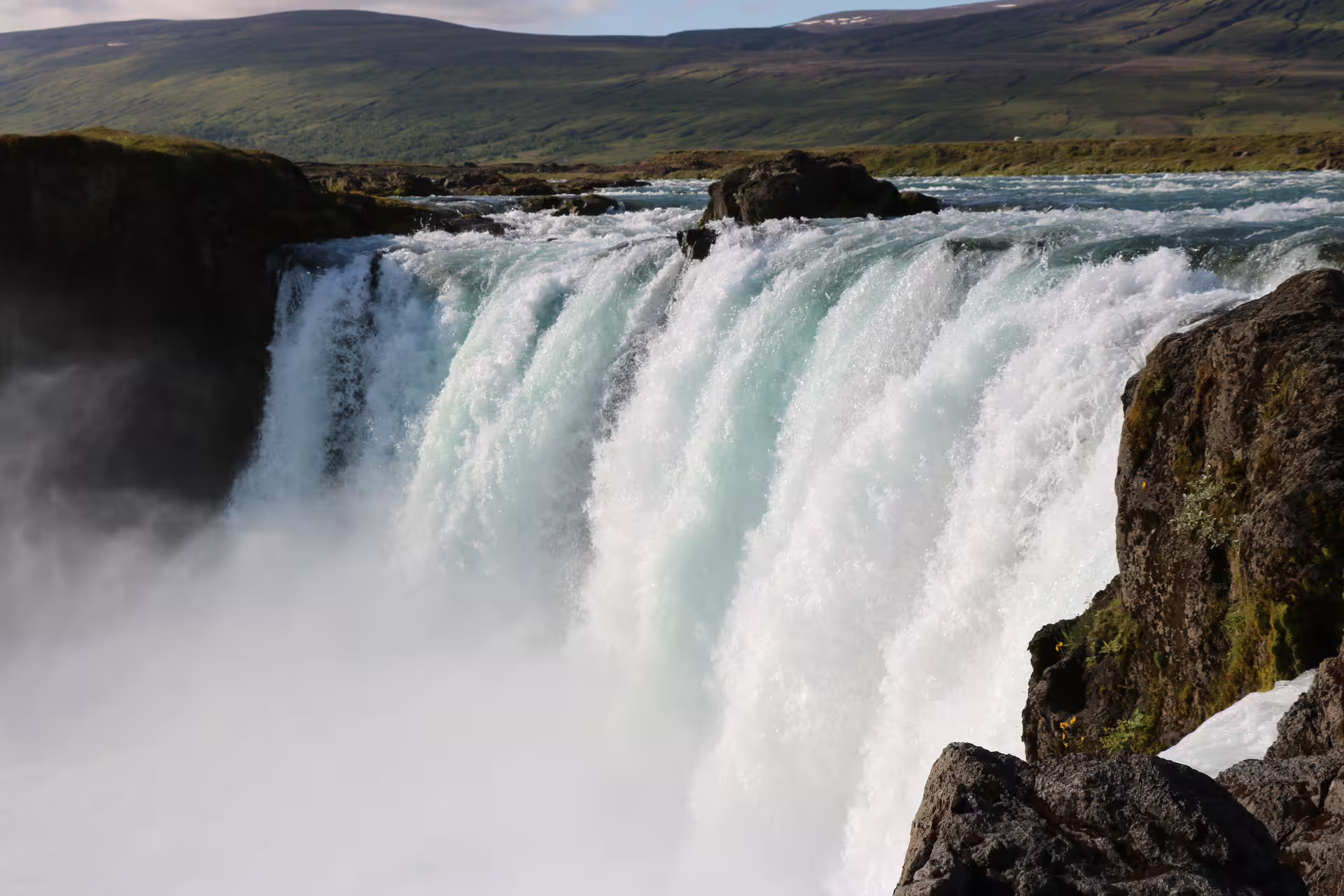 Godafoss waterfall cascading over rocky ledge with turquoise glacial water and green Icelandic hills in background