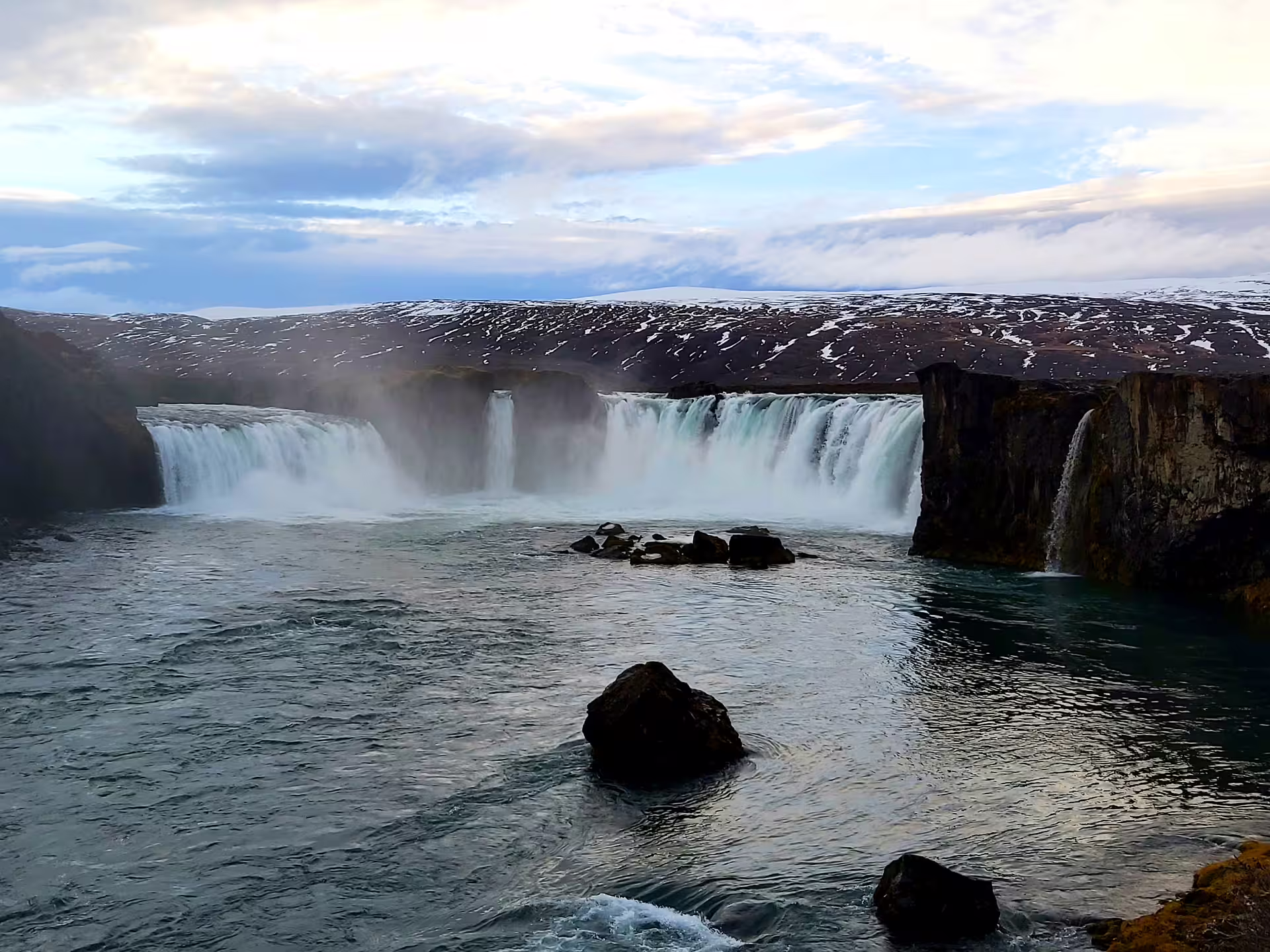 Wide cascading Godafoss waterfall with misty spray, rocky basin, and snow-dusted hills in North Iceland
