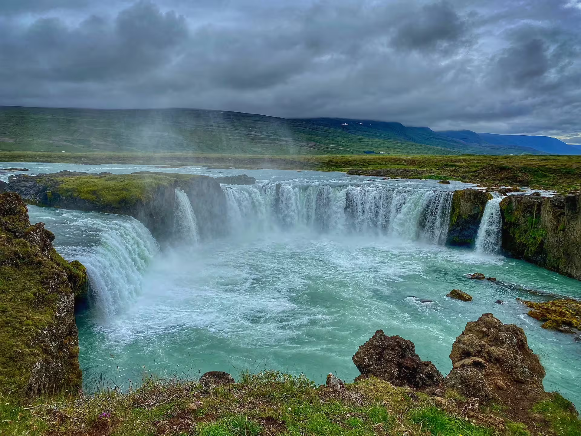 Godafoss waterfall with turquoise glacial waters and misty spray under dramatic cloudy skies in Iceland