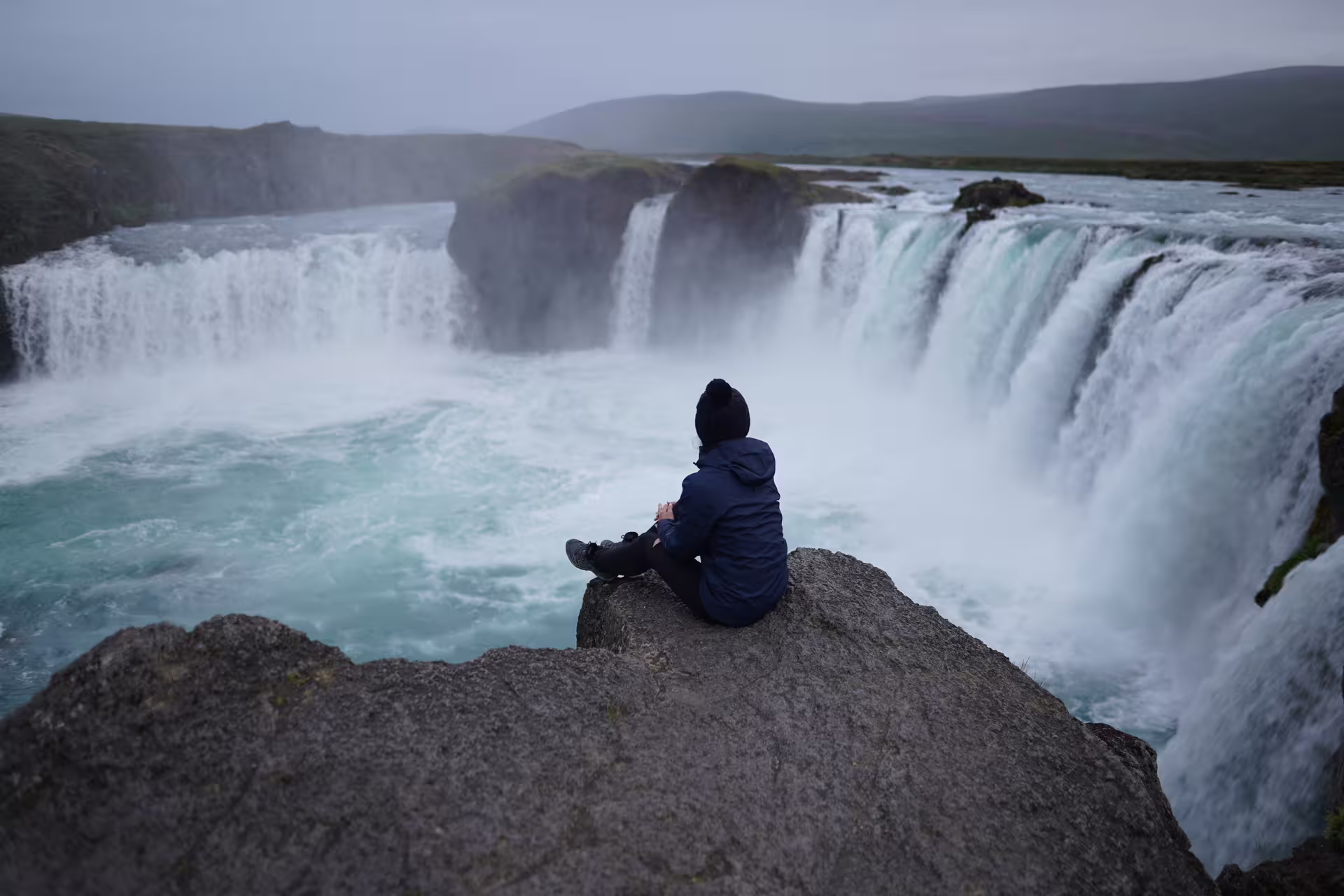 Person sitting on rock ledge overlooking Godafoss waterfall in Iceland on overcast day