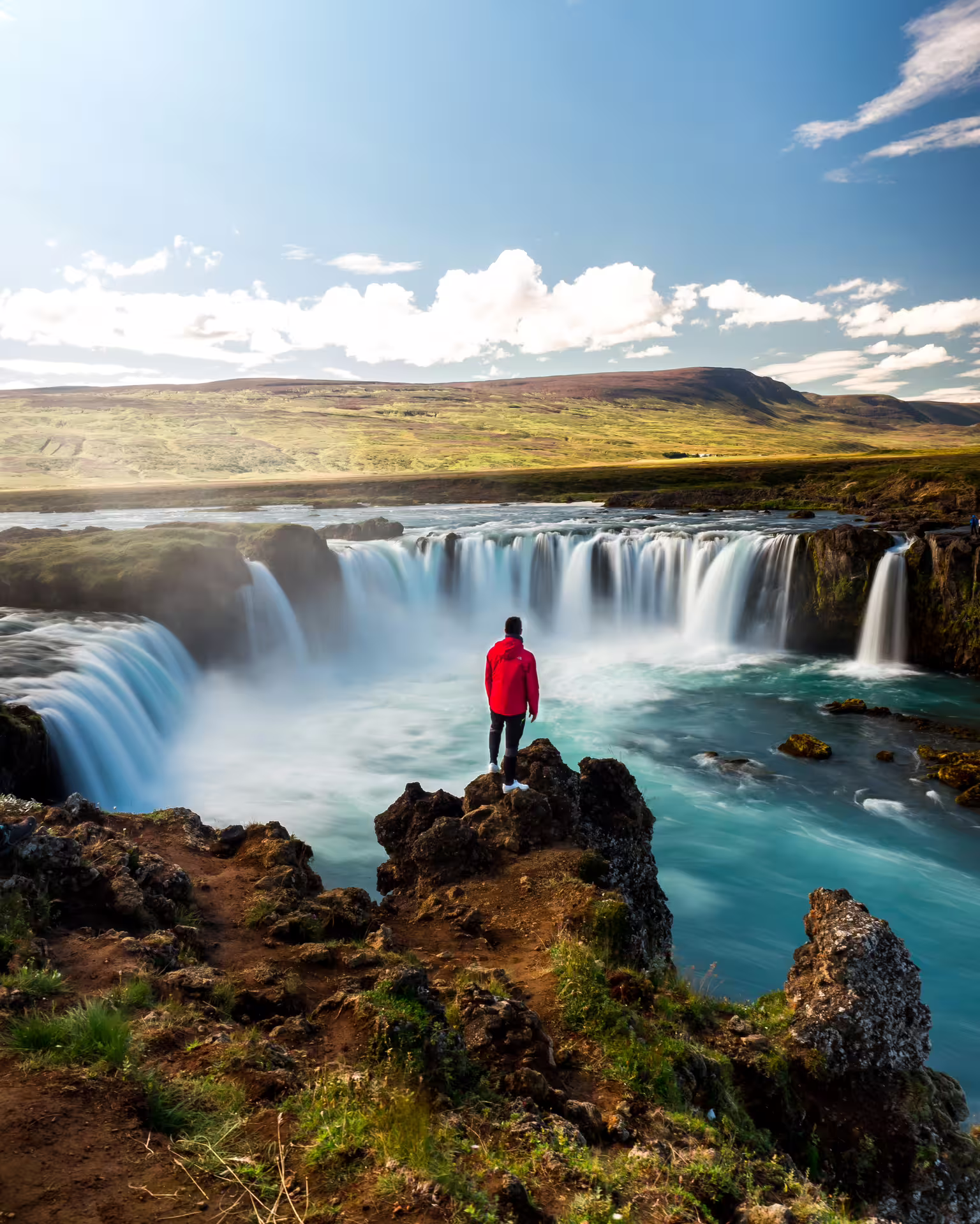 Person in red jacket standing on rocks overlooking Godafoss Waterfall with turquoise water in Iceland