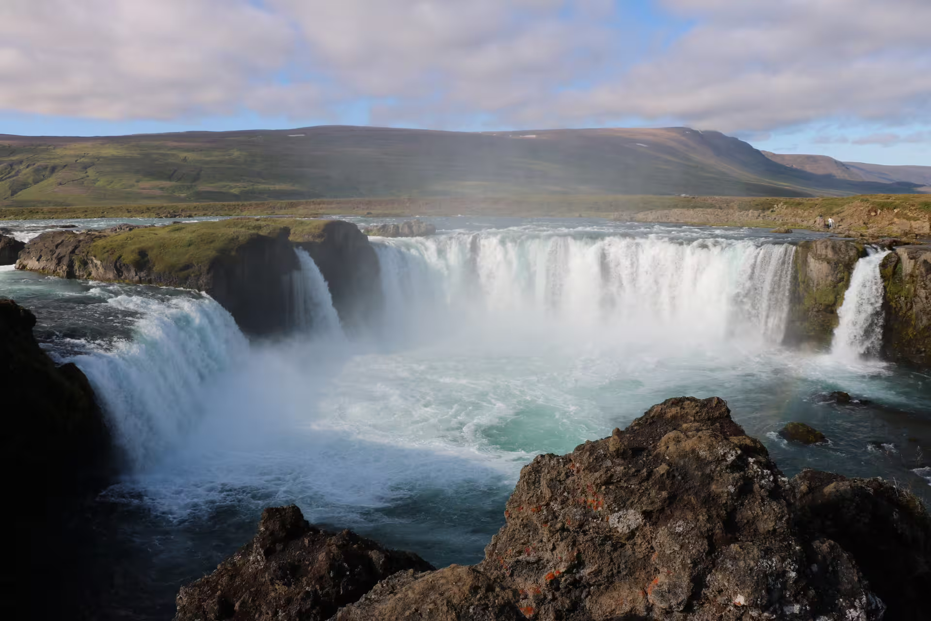 Godafoss waterfall cascading into turquoise pool with misty spray and green Icelandic highlands in background