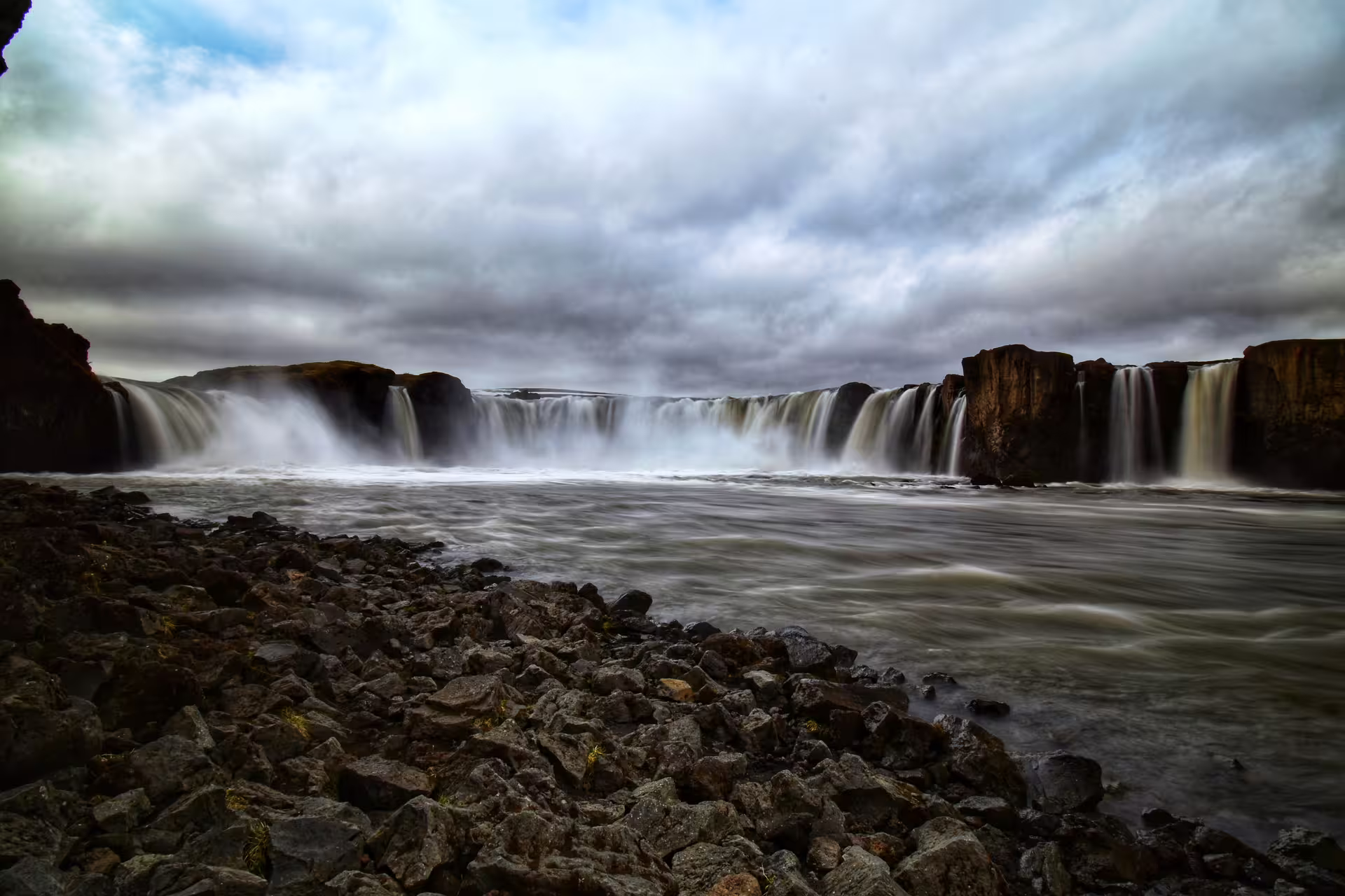 Godafoss waterfall cascading over basalt cliffs into misty river under dramatic cloudy sky, Iceland