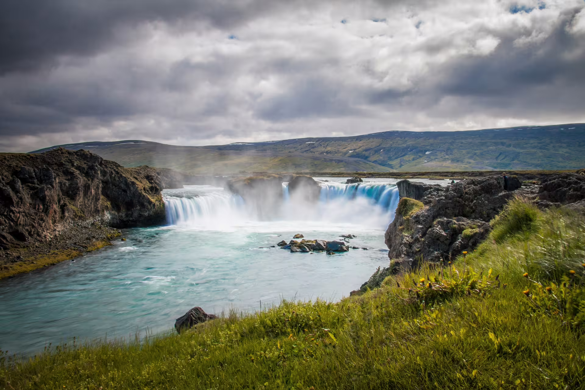 Godafoss waterfall with turquoise glacial water flowing over basalt rocks under dramatic cloudy skies in Iceland
