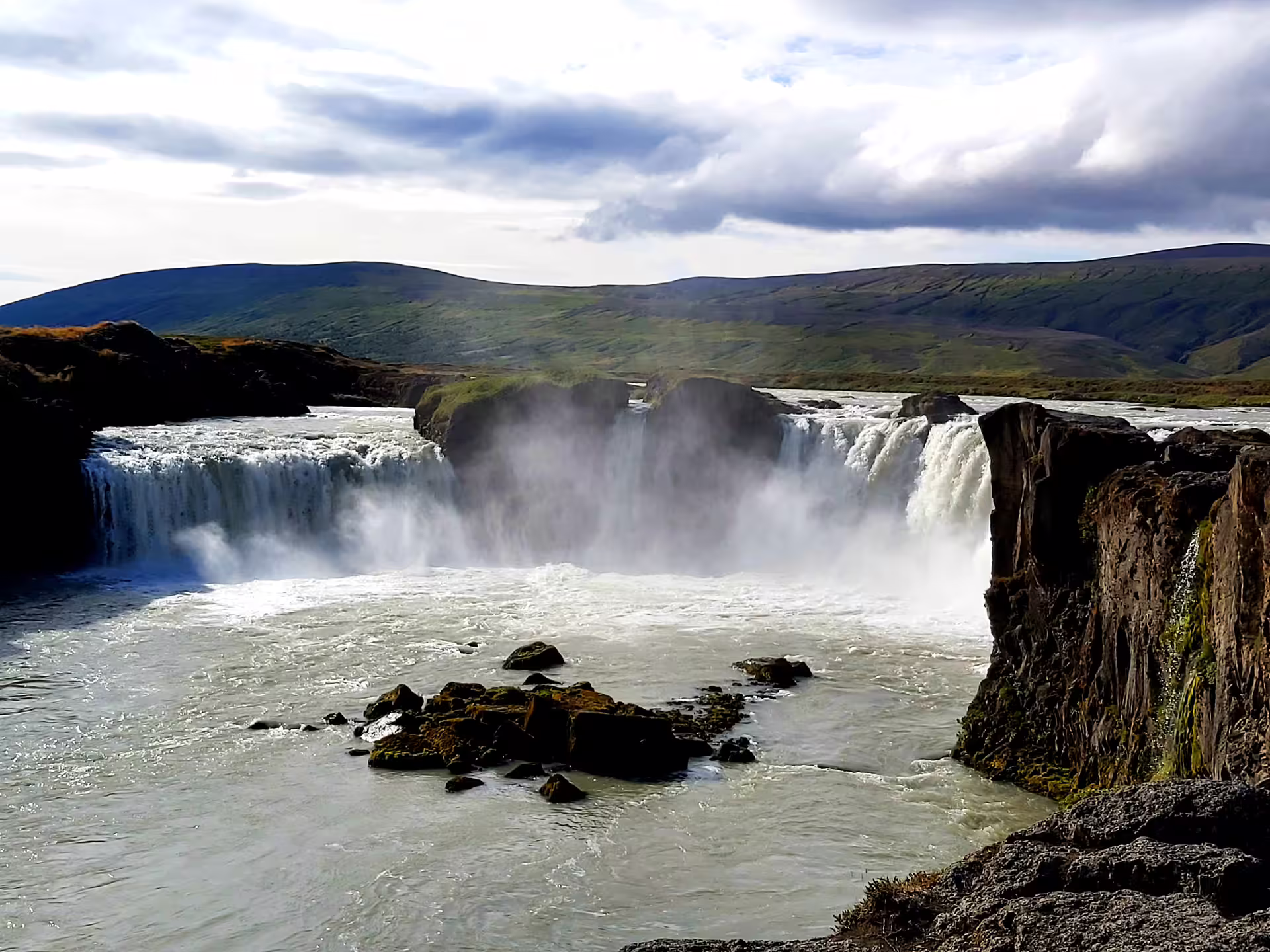 Powerful Godafoss waterfall cascading into misty pool with green hills in Iceland
