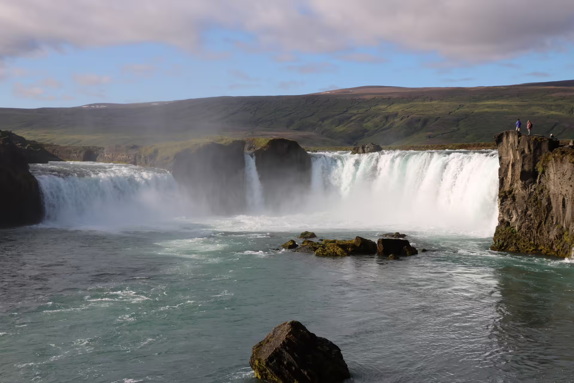 Godafoss waterfall cascading into turquoise river with misty spray and green Icelandic hills beyond