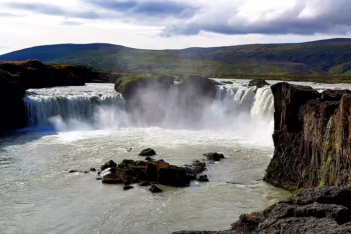 Godafoss waterfall cascading into misty pool with rocky landscape and green hills in North Iceland