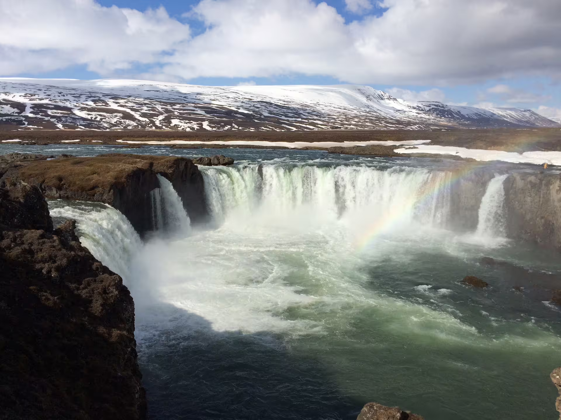 Godafoss waterfall in Iceland with rainbow mist and snow-capped mountains in background