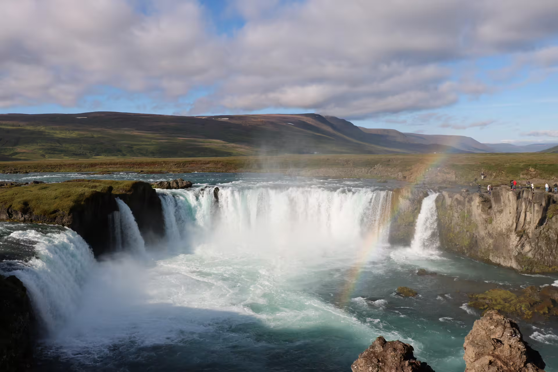 Godafoss waterfall in Iceland with rainbow mist and turquoise waters beneath cloudy skies