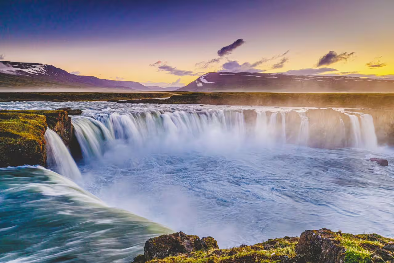 Godafoss waterfall cascading at sunset with snow-capped mountains in background, northern Iceland