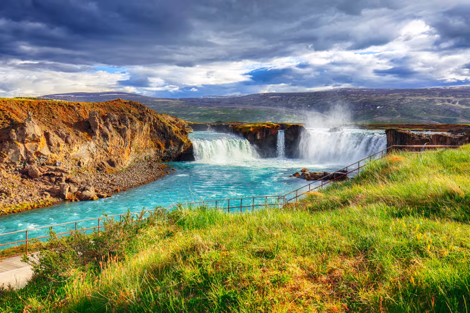 Godafoss waterfall with turquoise glacial river, lush green grass, and dramatic cloudy sky in Akureyri, Iceland