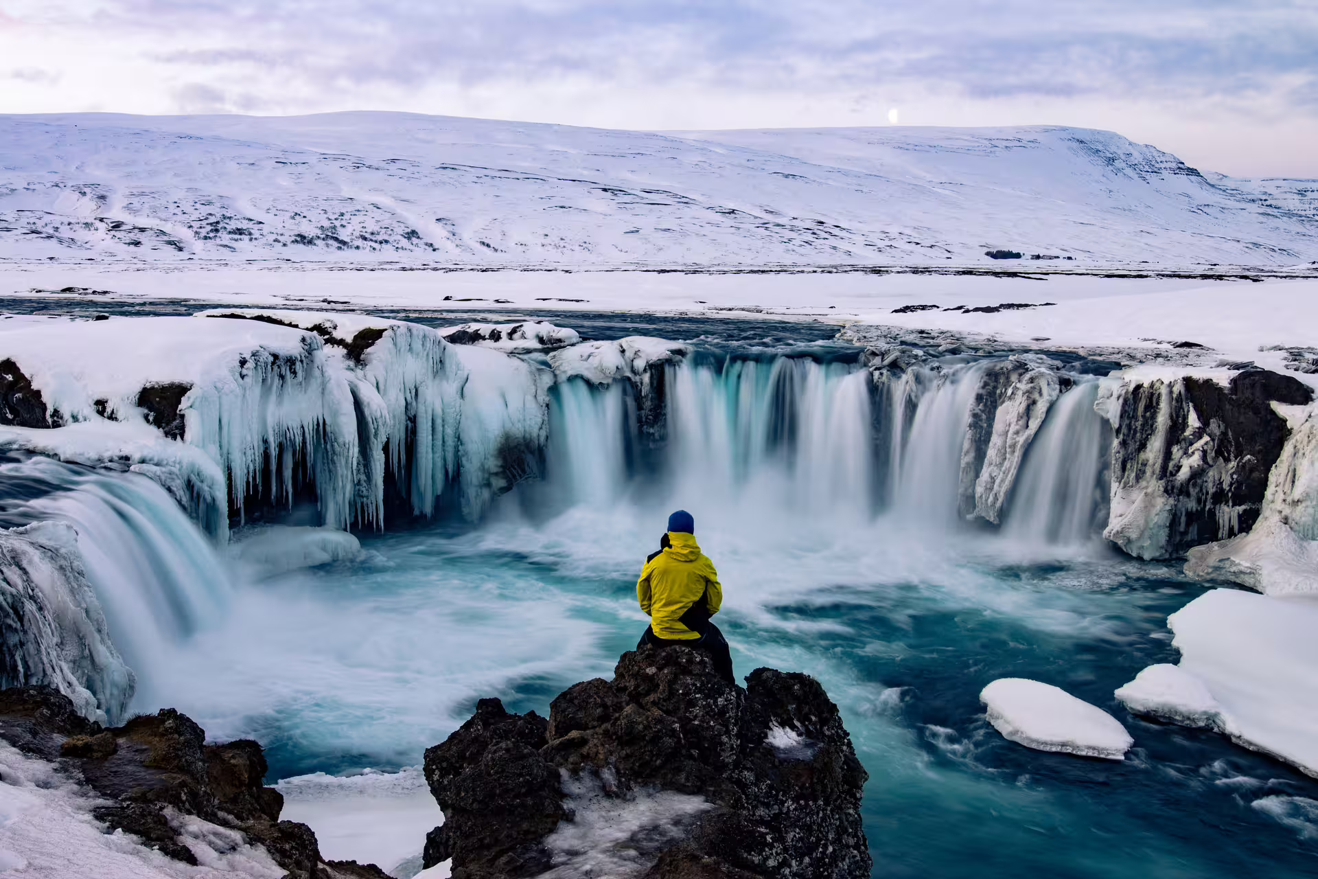 Person in yellow jacket sitting on rocks overlooking frozen Godafoss waterfall in winter, Iceland
