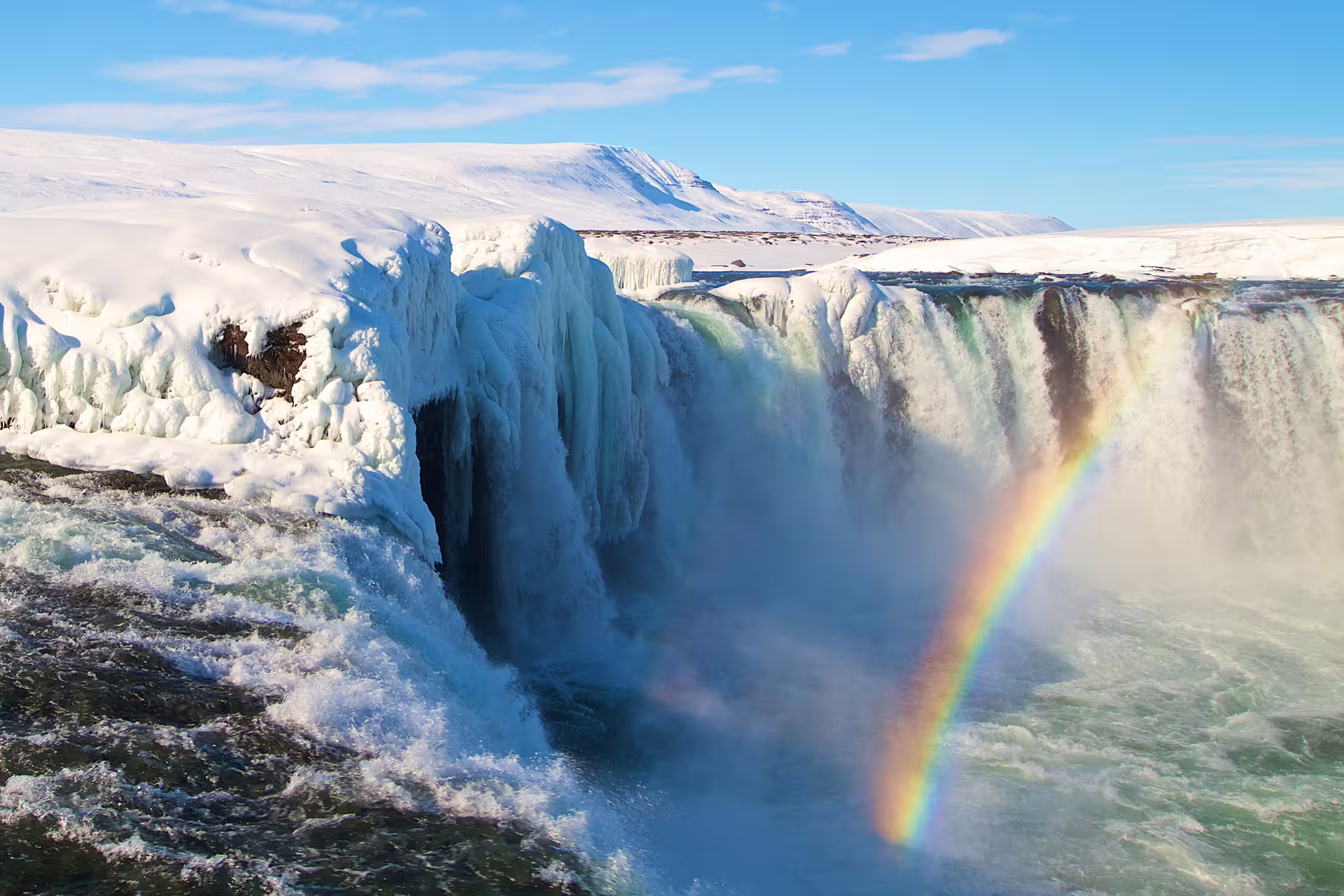 Godafoss waterfall in winter with ice formations and rainbow, snow-covered landscape in background