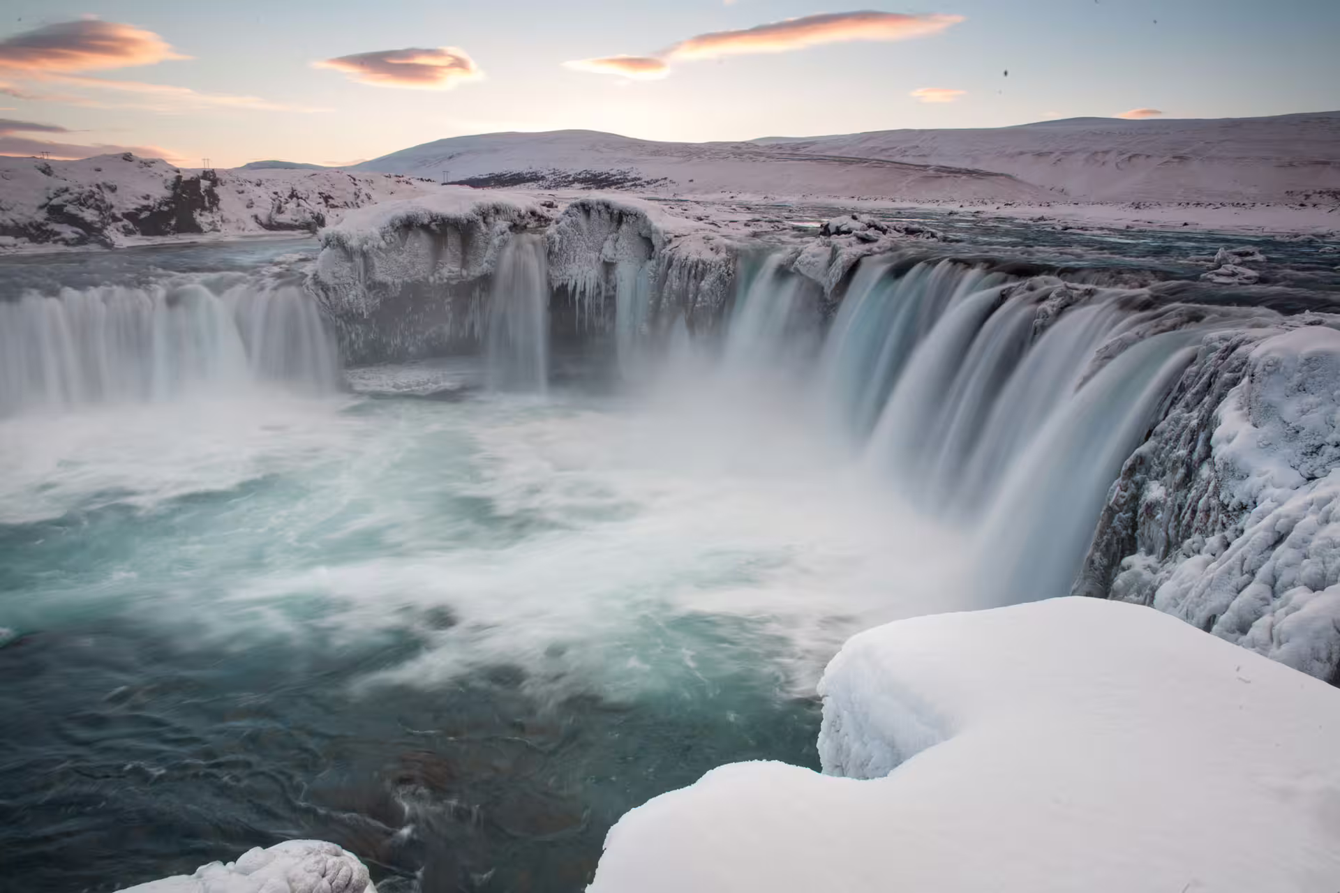 Godafoss waterfall in winter with snow-covered rocks, icicles, and turquoise rushing water at sunset, Iceland