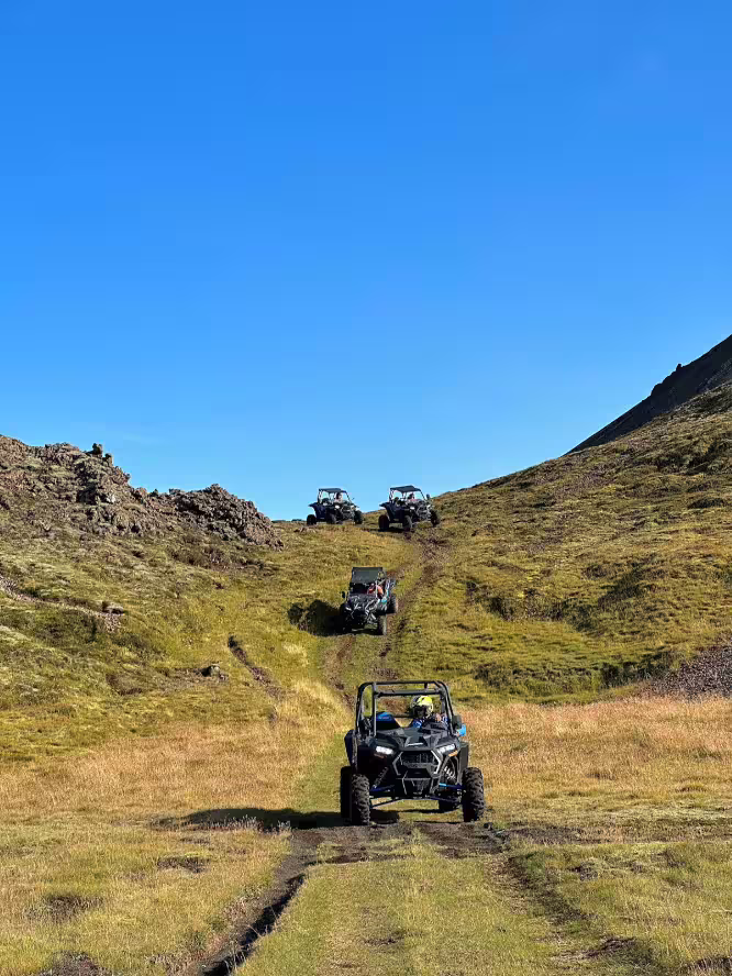 Three buggies driving in a convoy up a grassy hillside trail under a clear blue sky in Iceland