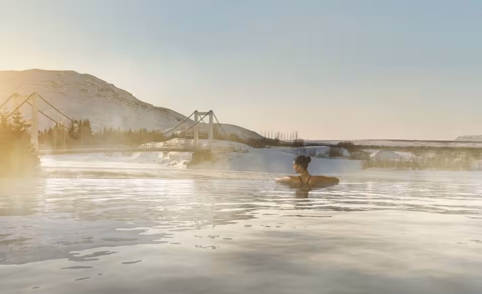 Woman relaxing in steaming geothermal lagoon with snowy mountains and bridge in background