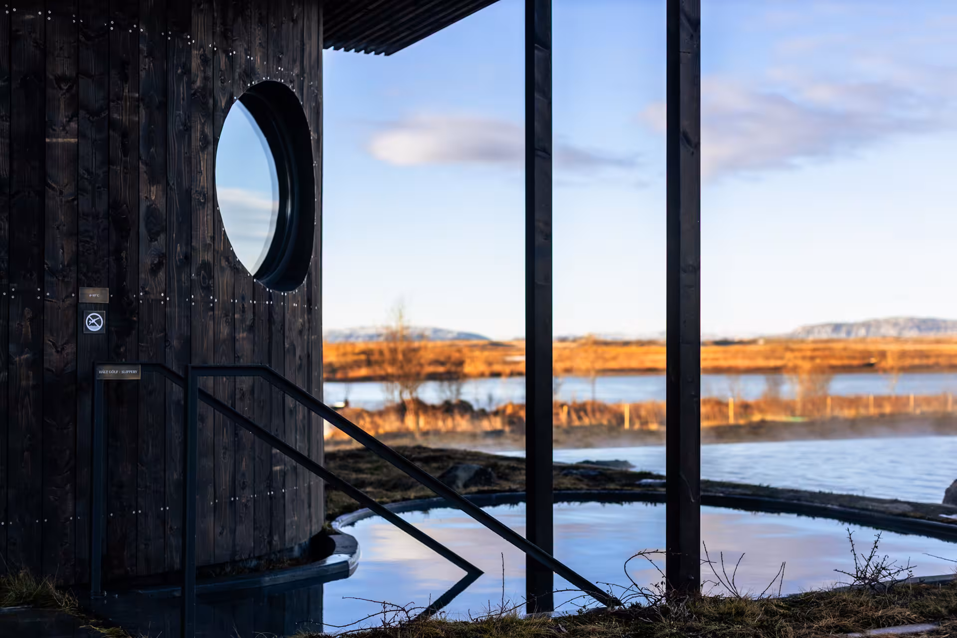 Dark wood building with circular window beside a steaming geothermal lagoon overlooking a golden Icelandic landscape