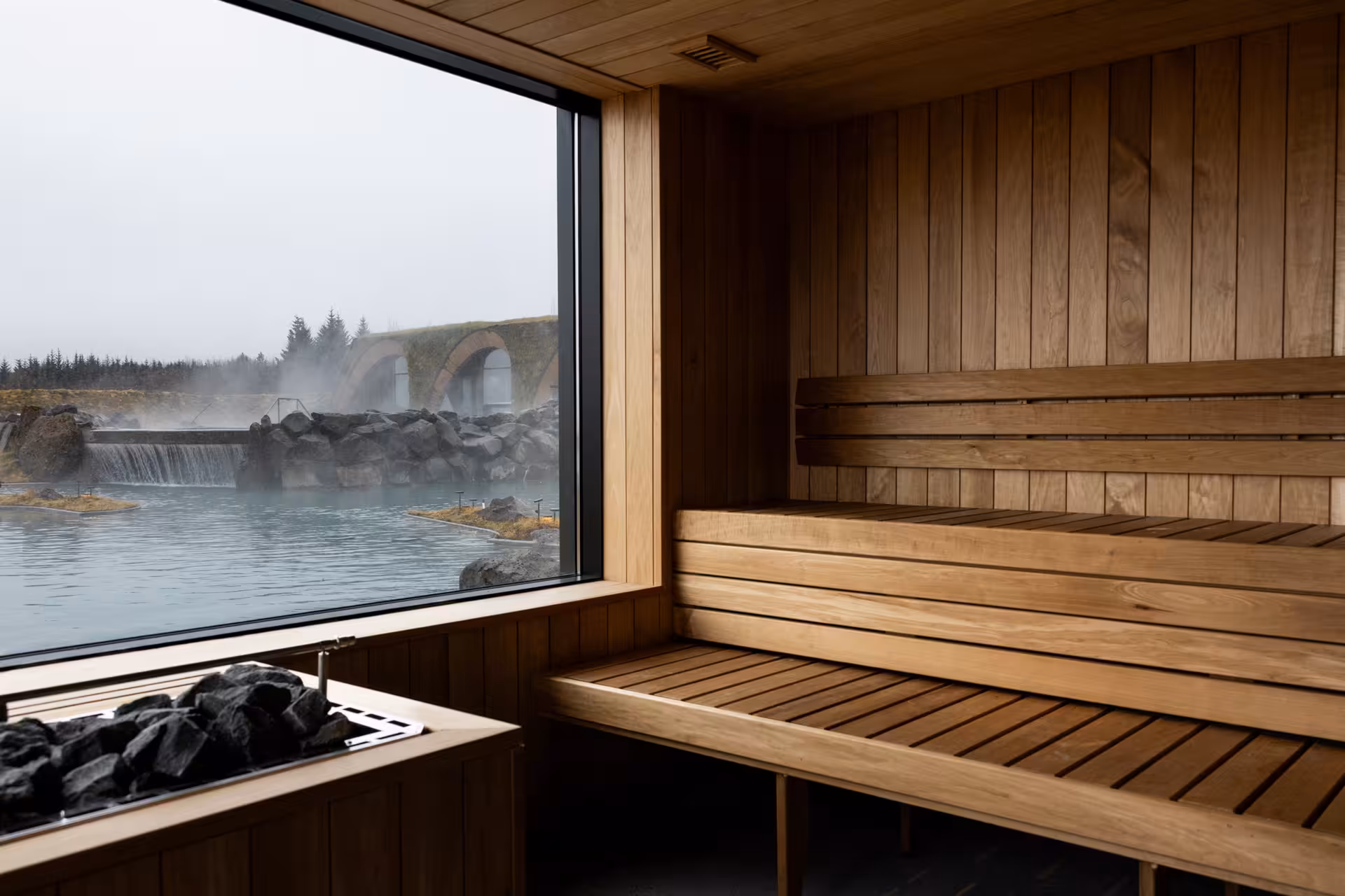 Wooden sauna interior with large window overlooking steaming geothermal lagoon and stone arches in Iceland