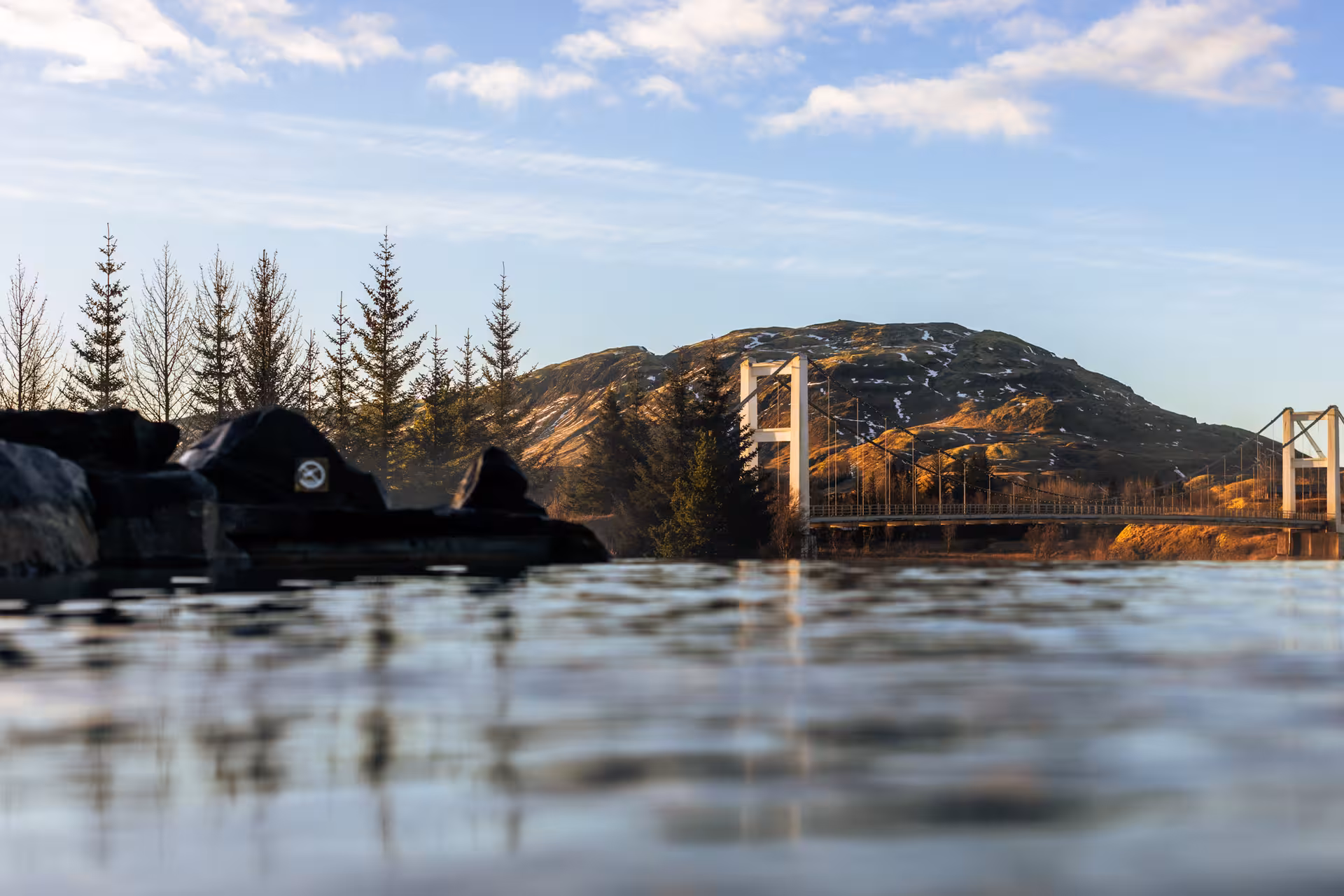 Geothermal lagoon with steaming water, suspension bridge, and snow-dusted hill in Iceland's Golden Circle