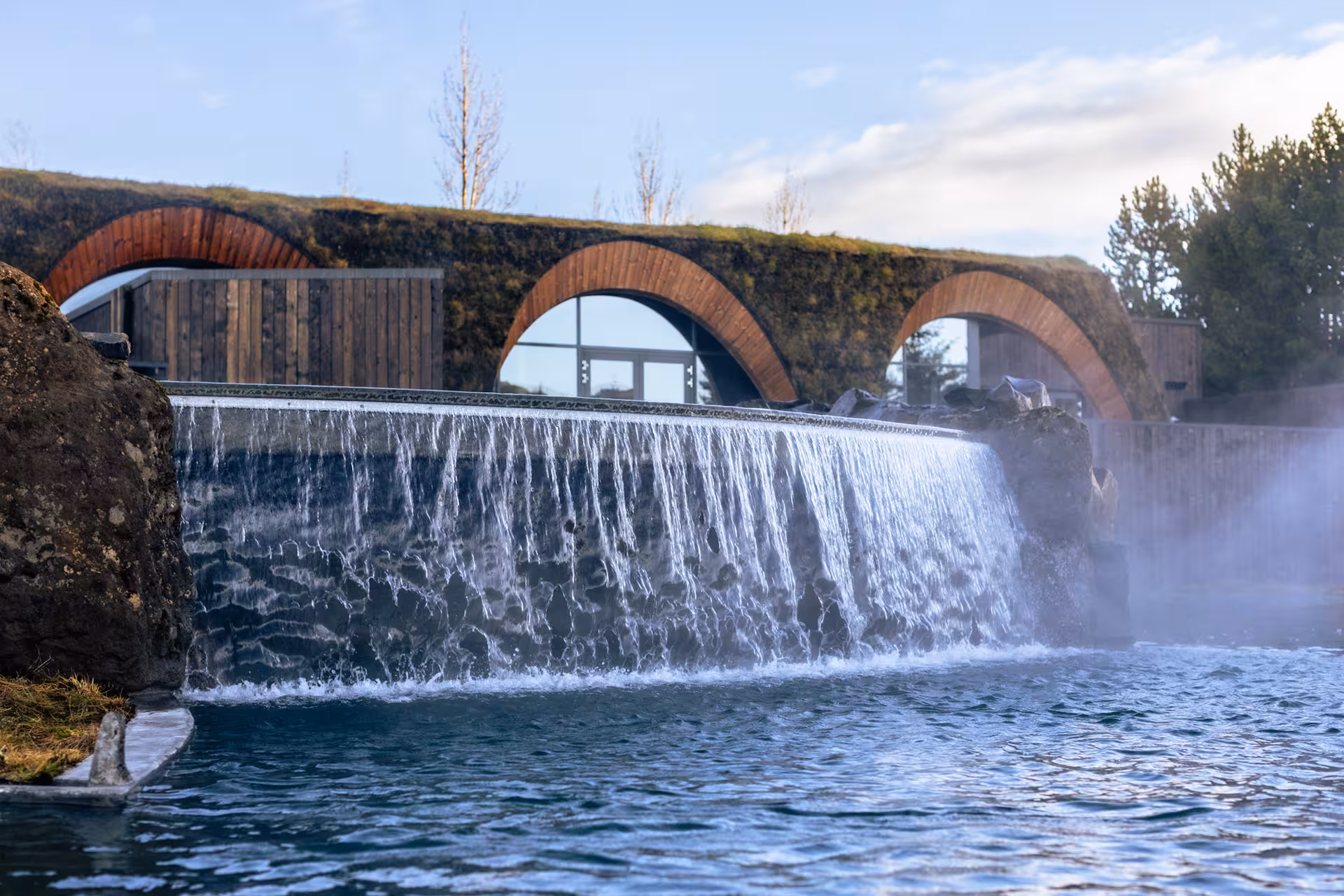 Steaming geothermal lagoon with cascading waterfall and arched wooden building with grass roof in Iceland