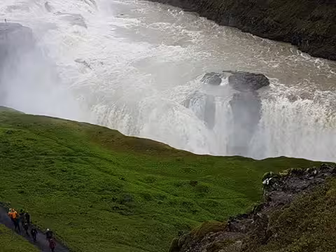Tourists walking along green hillside path overlooking powerful Gullfoss waterfall on Golden Circle tour