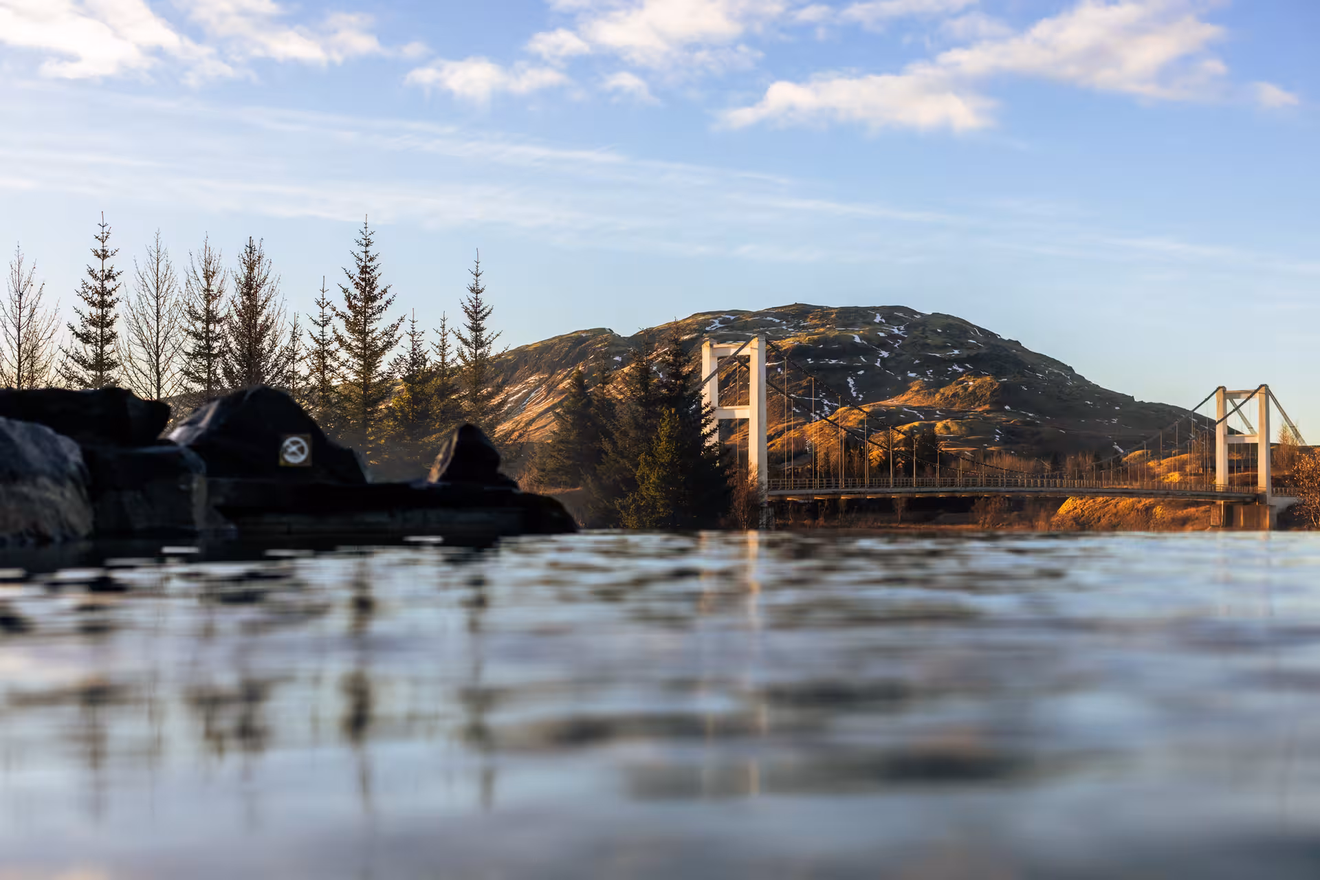 Suspension bridge over calm lake with snow-dusted volcanic mountain in Golden Circle, Iceland