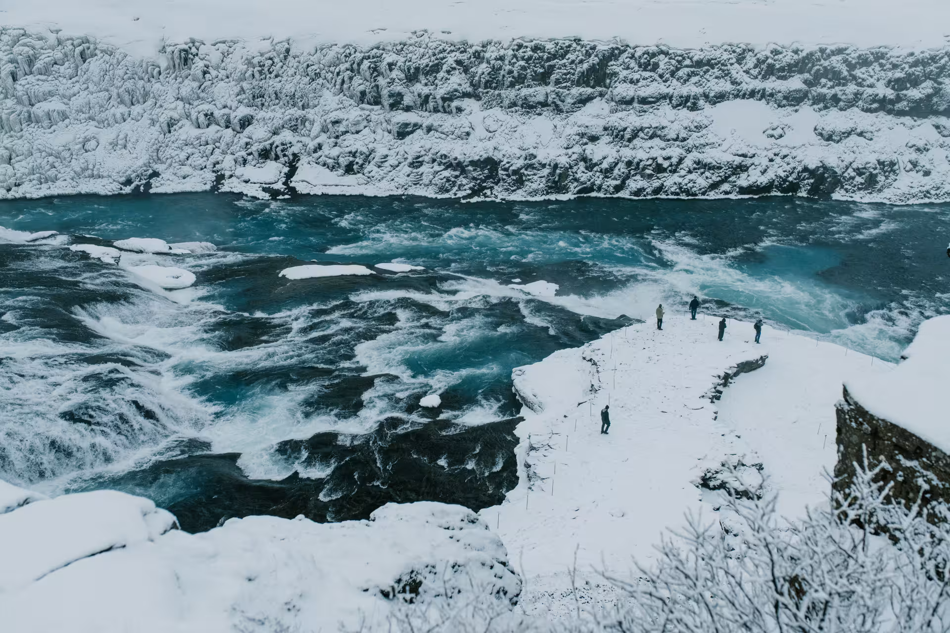 Visitors standing on snowy cliff overlooking turbulent glacial river rapids in Iceland winter landscape