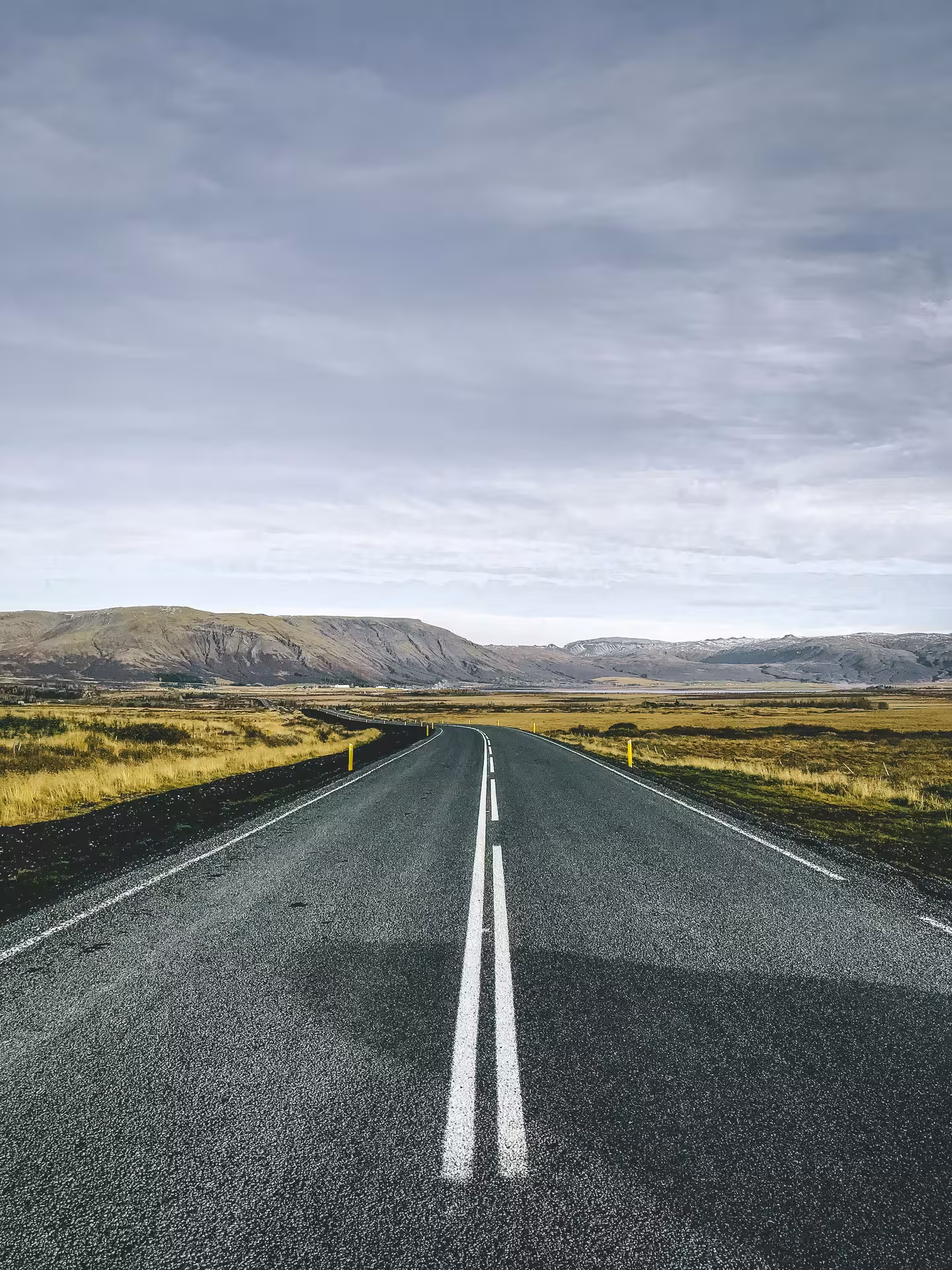 Empty two-lane road stretching through golden Icelandic highlands toward volcanic mountains under cloudy sky