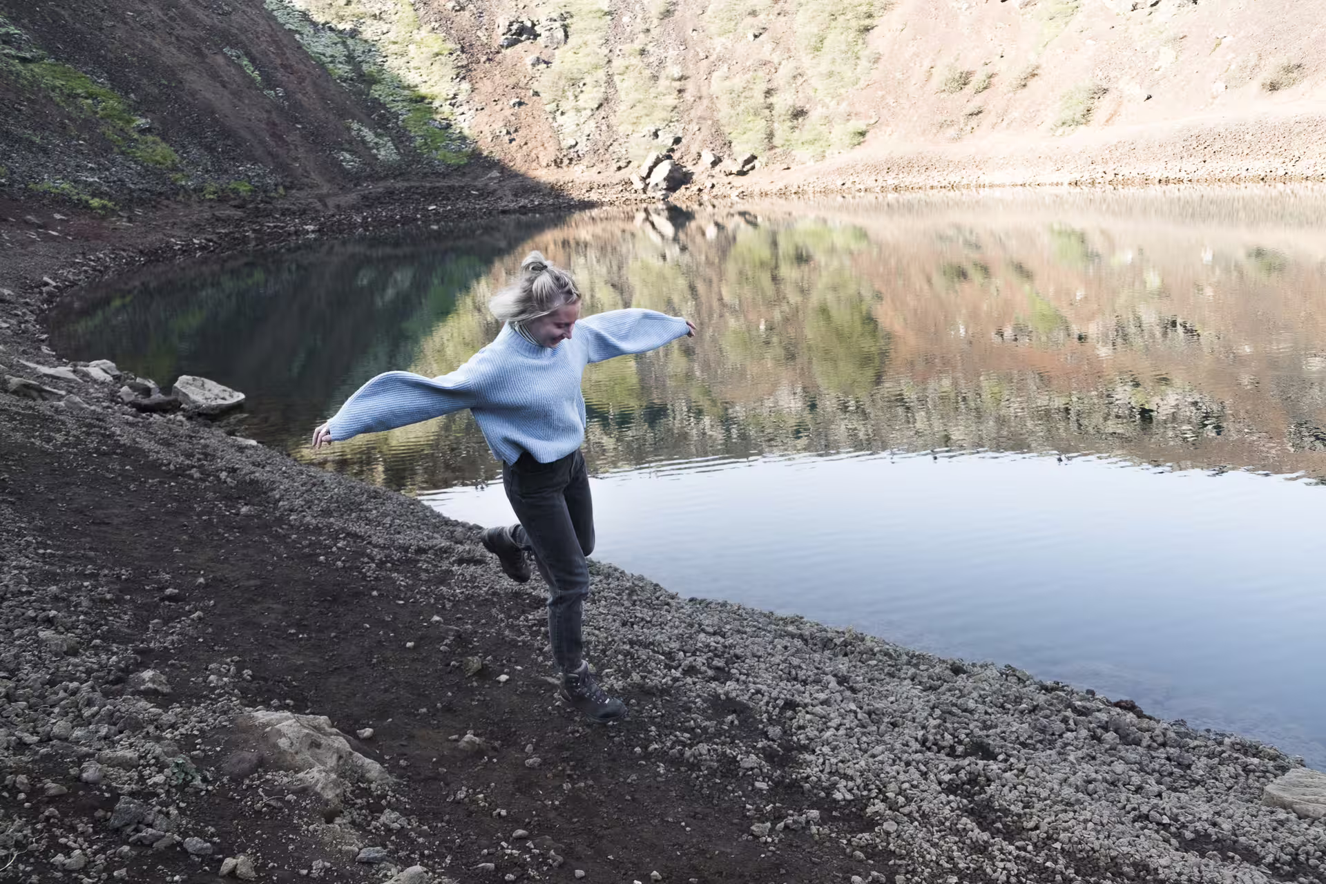 Woman balancing on rocky volcanic shore beside a calm reflective crater lake in Iceland