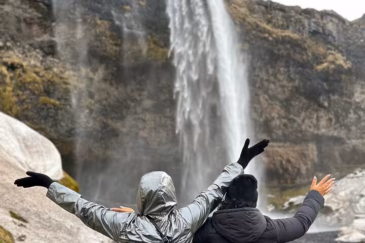 Two travelers with arms raised celebrating in front of a majestic Icelandic waterfall on a cloudy day