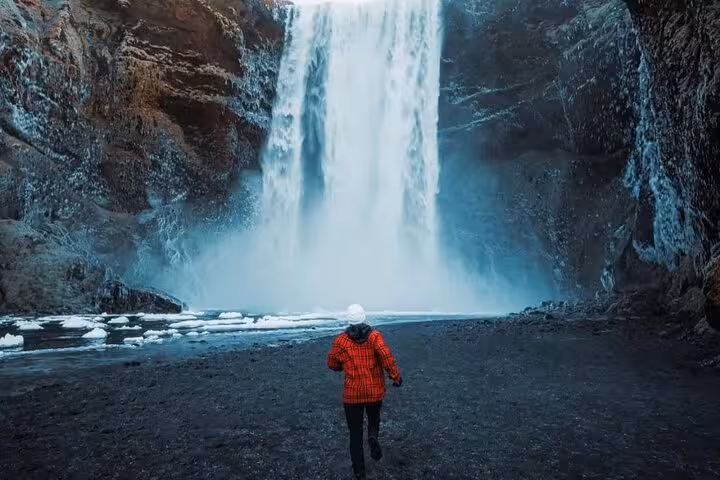 Person in red jacket walking toward large waterfall with icy rocks in Iceland