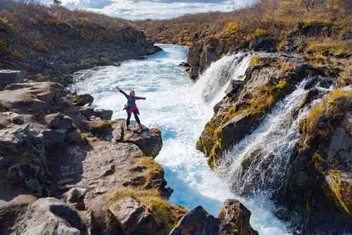 Hiker standing on rocky cliff with arms spread wide beside turquoise waterfall in Iceland