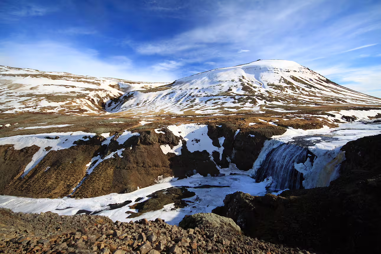 Snow-dusted volcanic canyon with waterfall and mountain peak under blue sky in Iceland