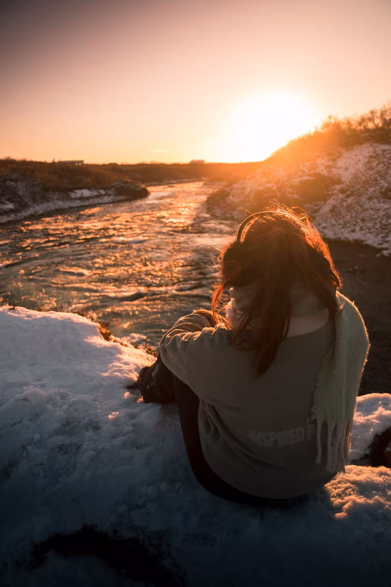 Woman sitting by a partially frozen river in Iceland during a golden sunset in winter