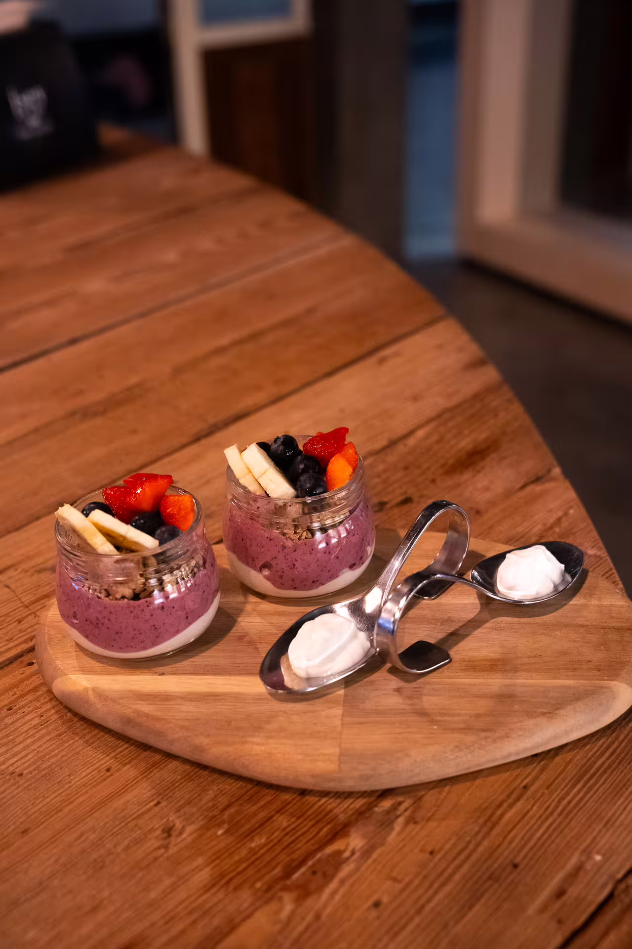 Two berry smoothie jars topped with strawberries, blueberries and banana slices on a wooden board with spoons