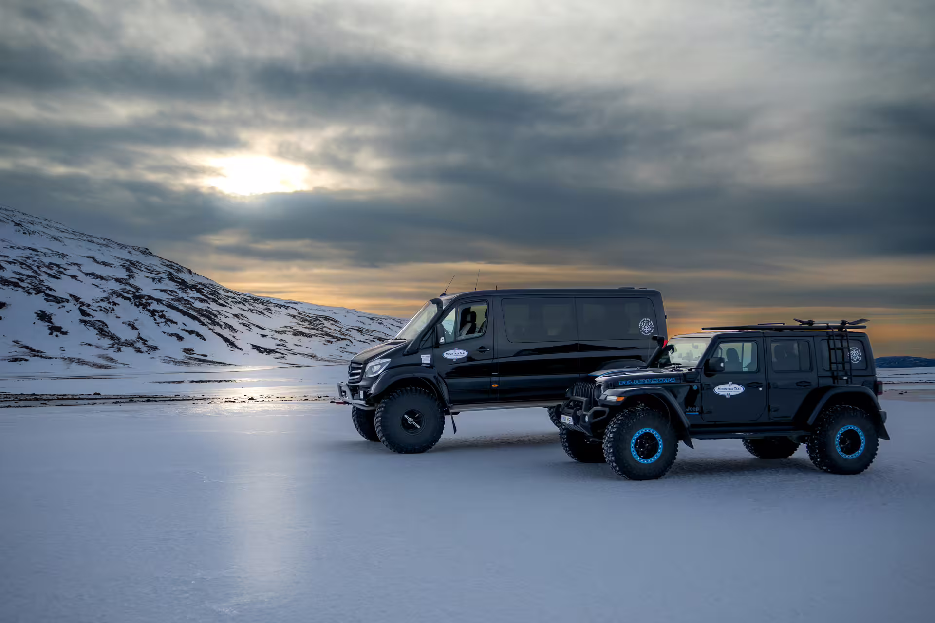 Two black lifted 4x4 vehicles on snowy Icelandic landscape with dramatic sunset clouds and snow-covered mountains