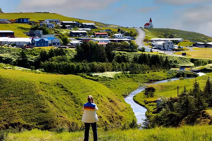 Person overlooking a scenic Icelandic village with a red-roofed church, green hills, and a winding stream